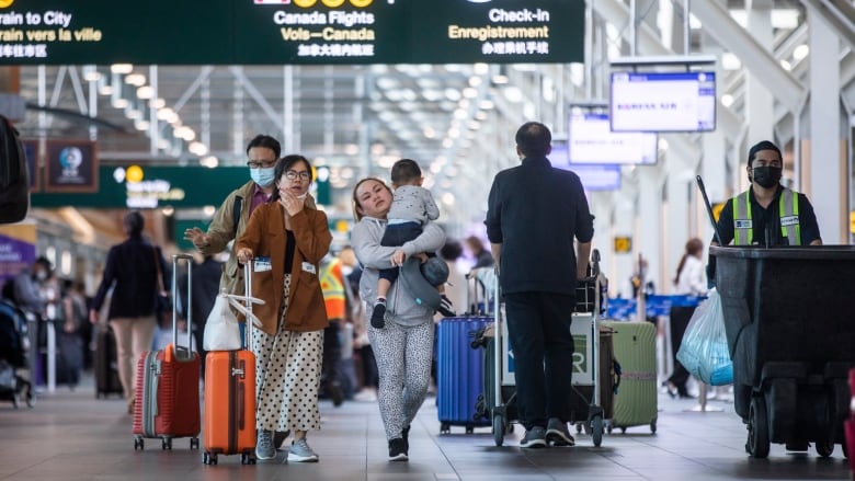 Travellers walk carrying and pushing luggage in an airport.