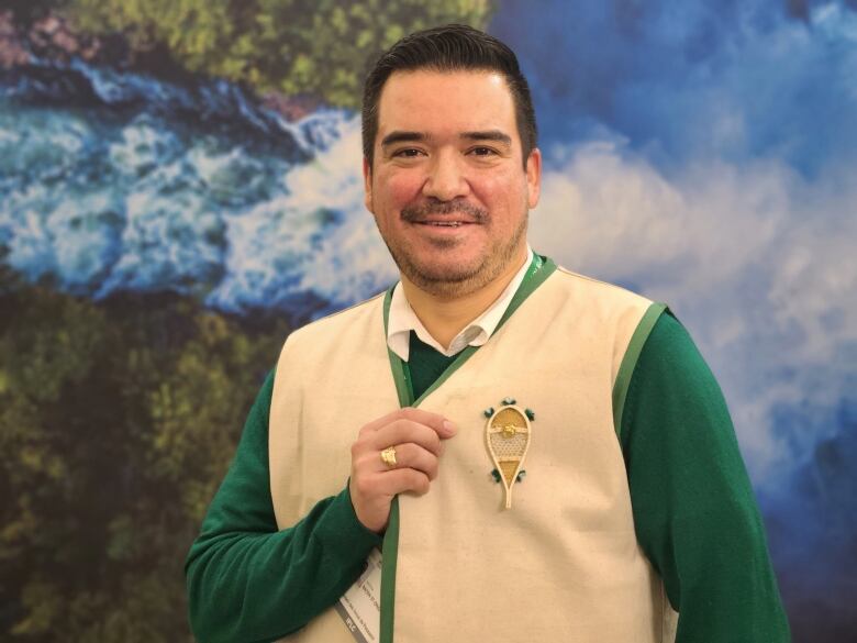 A man with dark brown hair is standing, looking at the camera. He is wearing a beige vest with green lining and a matching green shirt underneath. He is smiling and showing off a pin of a snowshoe on his vest.