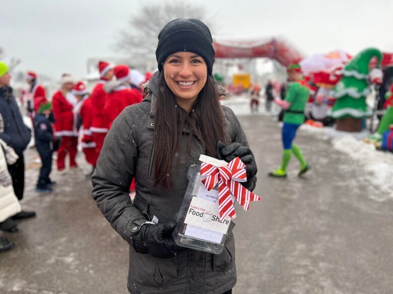 A woman in a black winter jacket smiles and holds a donation jar. Behind her, people in Santa and elf costumes are walking around. 