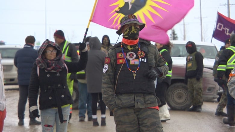 Protesters, some holding flags and others wearing reflective vests, stand outside.