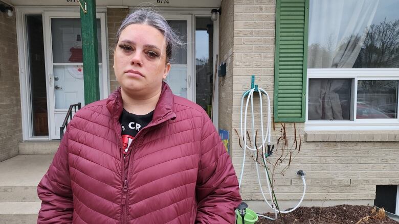 An upset woman with white and purple hair and nose piercings wearing a black t-shirt and a red jacket standing outside her front door