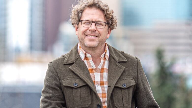 A man wearing glasses leans against a railing with a view of Calgary behind him.