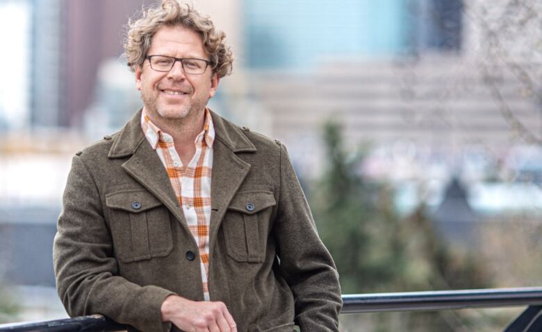 A man wearing glasses leans against a railing with a view of Calgary behind him.