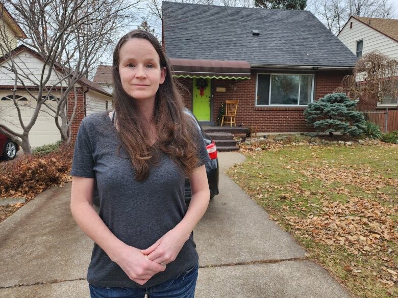 A woman with brown hair wearing a grey t-shirt and blue standing in front of her car on her driveway