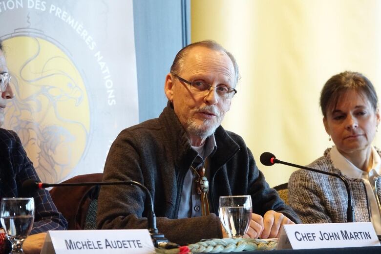 Man looks to his right as he is speaking into a microphone as part of a panel of speakers. 