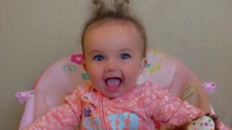 A baby with an open-mouth smile sits in a baby chair with a stuffed toy on her lap.