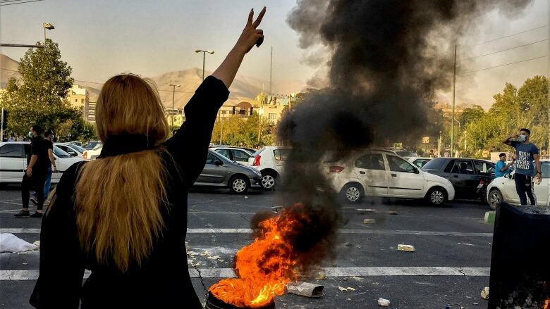 A woman with blond hair hanging down over her back, standing in front of a burning tire and a line of cars, raises her right hand in the air with two fingers in a V.