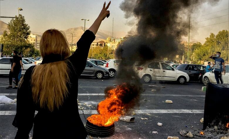 A woman with blond hair hanging down over her back, standing in front of a burning tire and a line of cars, raises her right hand in the air with two fingers in a V.