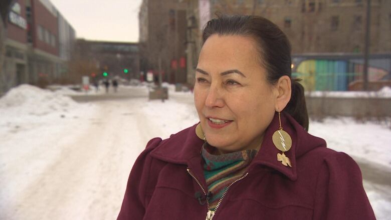 A woman wearing a maroon jacket and long gold earrings stands against a snowy backdrop. She has dark brown hair tied in a ponytail.