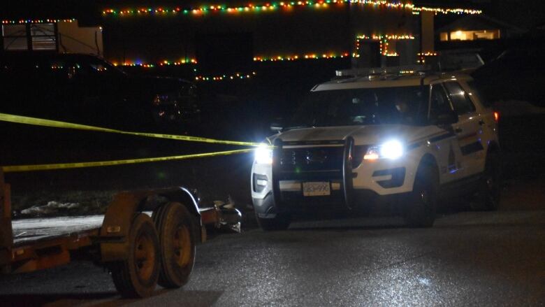 A white RCMP cruiser is parked on a dark suburban street in front of police tape. A house framed with Christmas lights can be seen in the background.