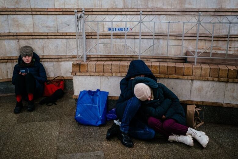 A woman sits on the ground in a subway station with a phone in her hand. Beside her is a couple, also sitting on the ground, hugging eachother 