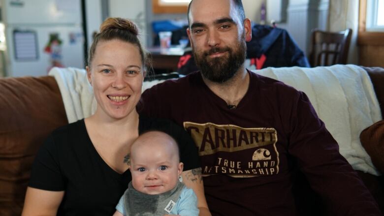 A woman smiles holding a wide-eyed baby on her lap. A man has his arm around the woman and is smiling. 