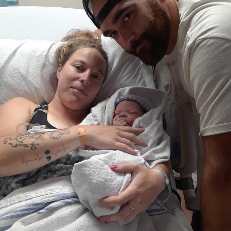 A woman is lying in a hospital bed holding a newborn baby adorned in a knit hat and hospital blanket. A man wearing a hat and white t-shirt leans into the frame. 