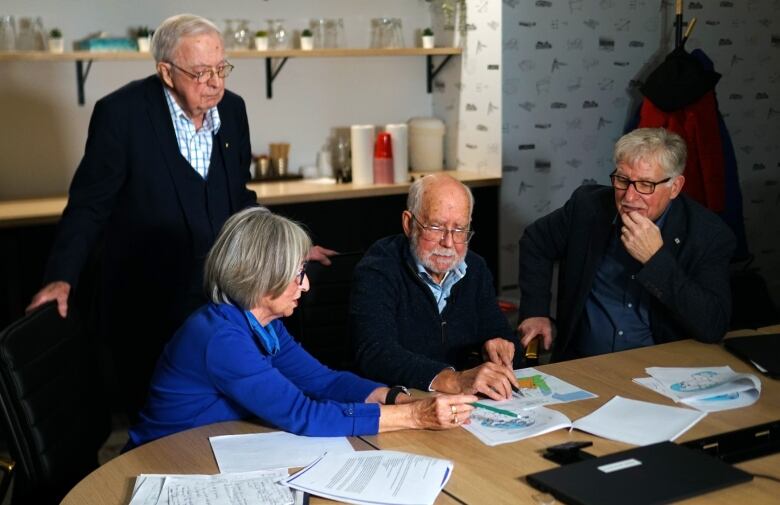 An older man wearing a cardigan stands over three people seated at a conference table as they look at a spread of papers. 