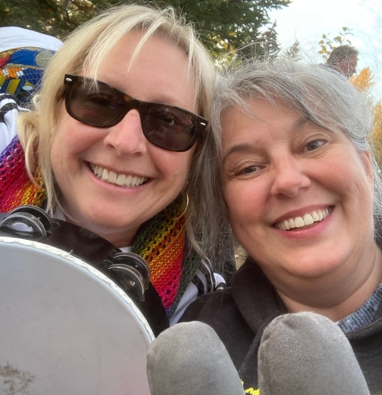 Two women take a selfie holding a tambourine and drum sticks outdoors.