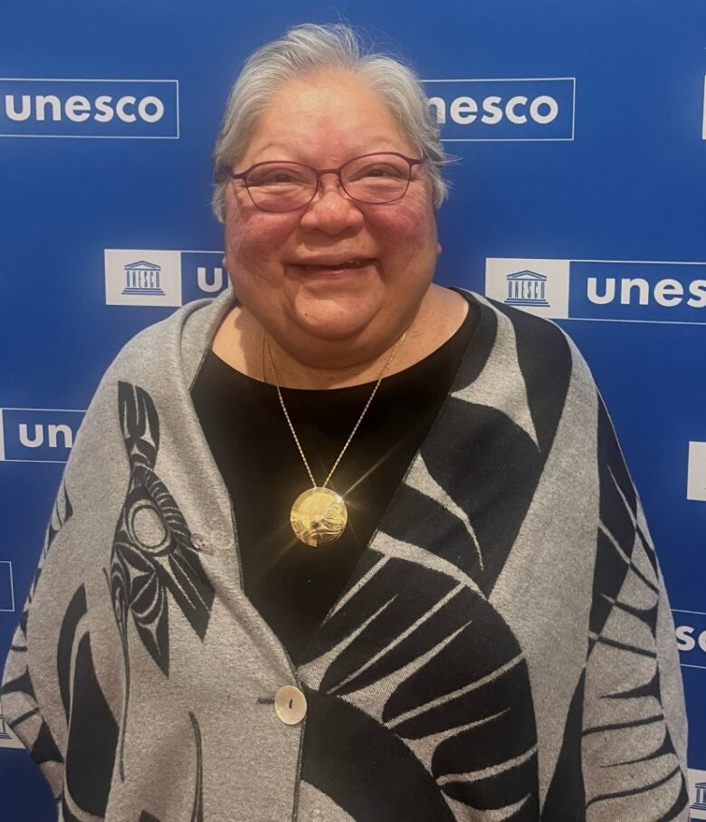 An Indigenous woman with glasses poses in front of the UNESCO blue backdrop. Shes wearing glasses, a bright smile and a  grey and black sweater.