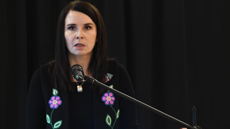 A woman with dark hair past her shoulders and wearing a black shirt with pink-and-blue beaded flowers speaks into a microphone.