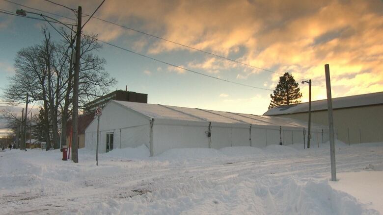 A large white tent is shown in a parking lot surrounded by snow as the sun sets in the background.