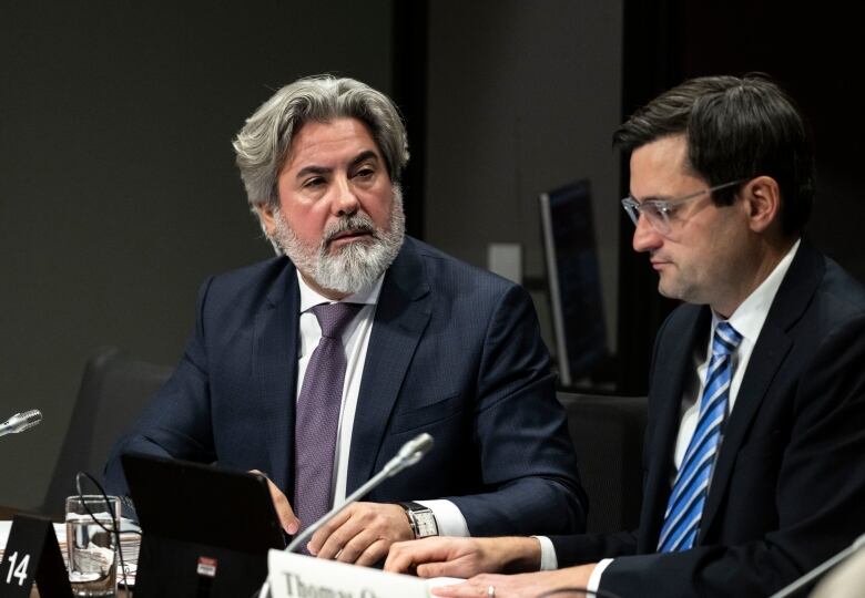 Two men in suits sit alongside each other at a conference table.