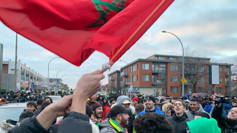 Morocco fans cheer and drum on Jean-Talon Avenue. A man waves a Moroccan flag overhead.