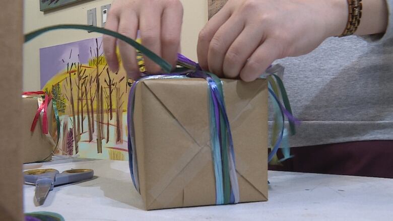 A close up shot of hands wrapping a present with brown paper and coloured ribbons, with a  pair of scissors beside the box.