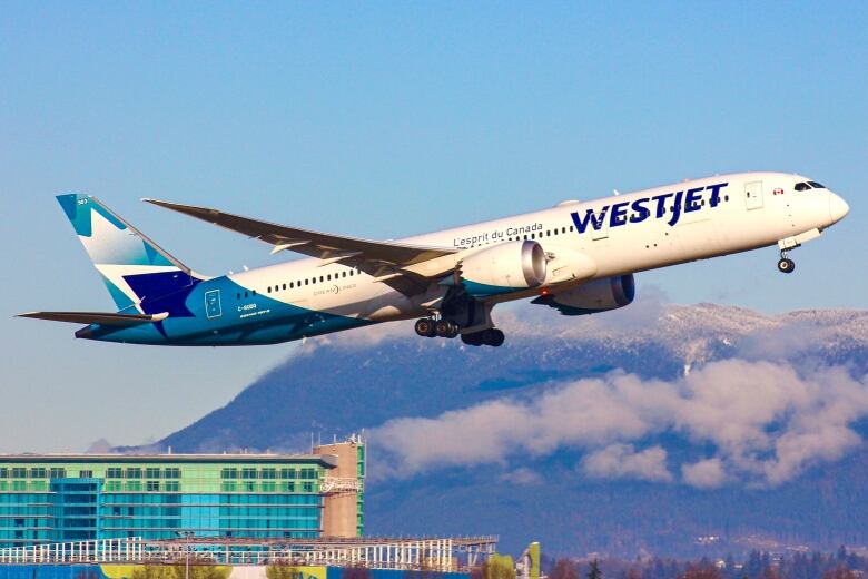 Blue and white WestJet plane is mid-air taking off from YVR airport. In the background is the YVR Fairmont hotel and snowcapped mountains covered in clouds. 