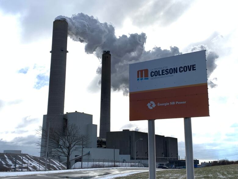 A red and white sign with black letters stands in front of an indsutrial site with billowing smoke stacks.