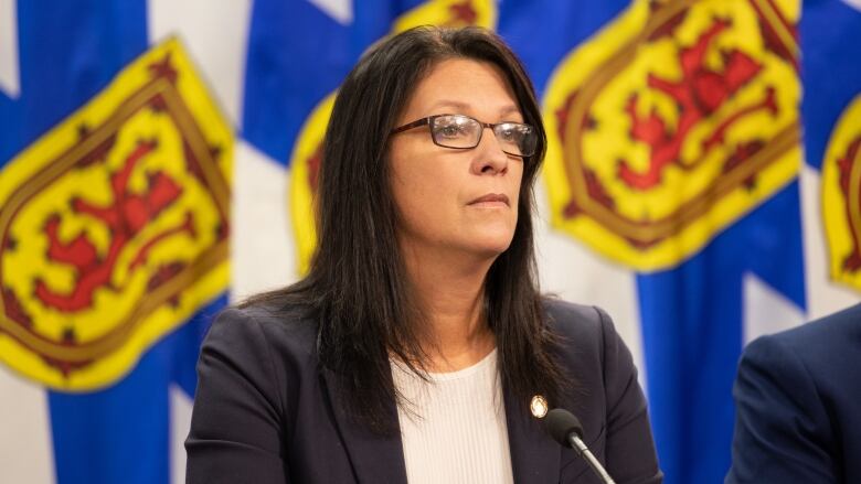 A woman with long black hair and glasses sits at a table and listens to questions.