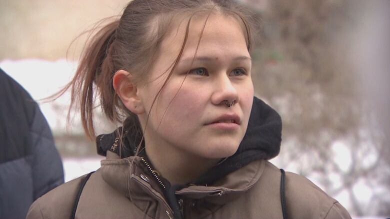 A teenage girl with her hair pulled back stands outside and looks at someone.