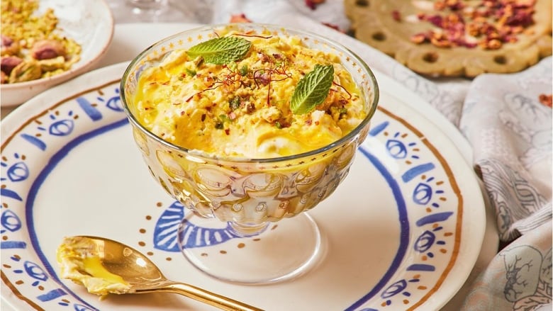 A footed glass bowl with a yogurt dessert in it. It's on a blue and white plate with a gold spoon next to it, and sitting on top of a table with a patterned tablecloth.