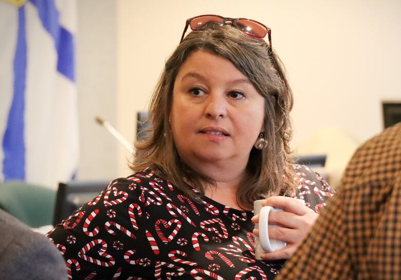 A woman with a candy-cane-print blouse and glasses on her head holds a coffee cup.