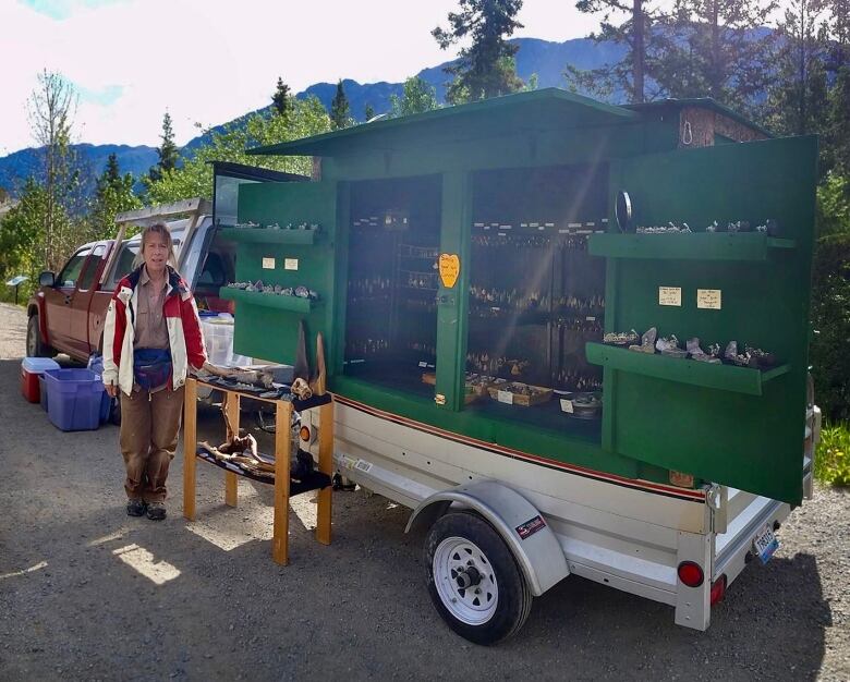 A woman stands outside, beside a vehicle trailer opened to display jewelry. 