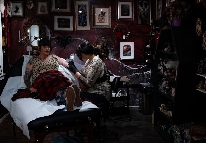 A woman tattoos another woman in a dark, gothic-inspired tattoo studio.