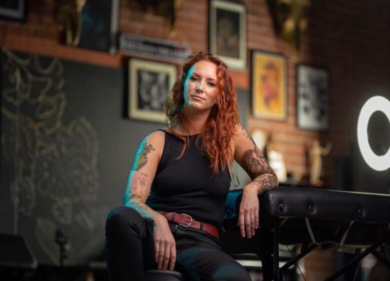 A red-haired woman bearing her tattoo-covered arms sits leaning against a tattoo table in a parlour whose walls are covered in framed art.