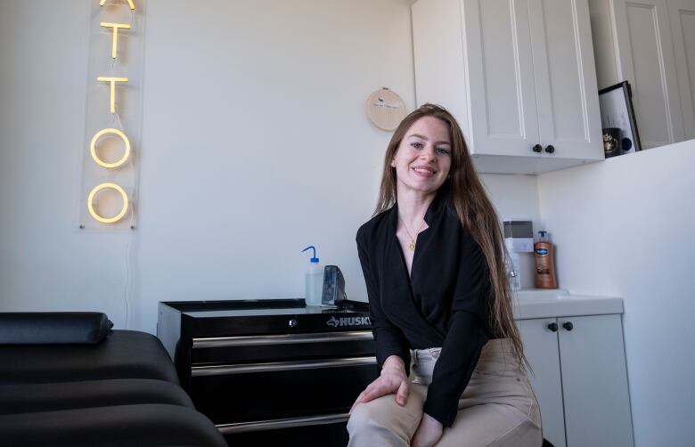 A young woman wearing a black blouse and beige trousers sits in a minimally decorated white room next to a black tattoo table. Yellow neon letters spelling 'tattoo' are attached in a vertical line on the wall.