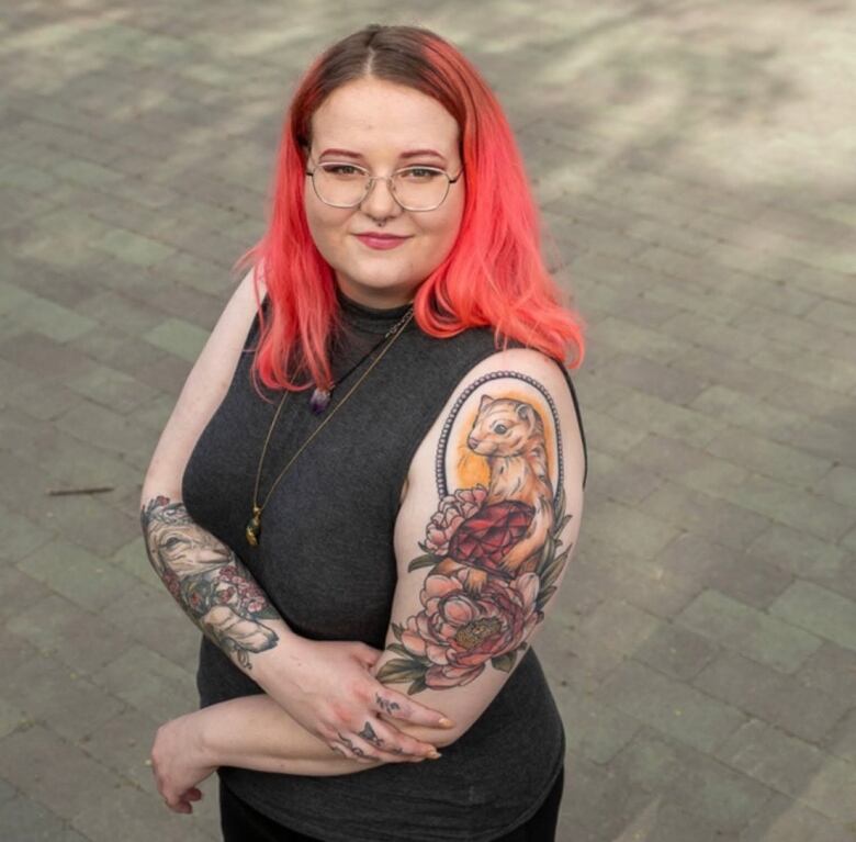 A person with long, coral hair stands with their tattoo-covered arms folded over their stomach.