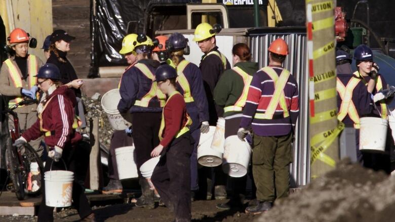 About a dozen people in hardhats and reflective work vests carry buckets amid dug-up earth.