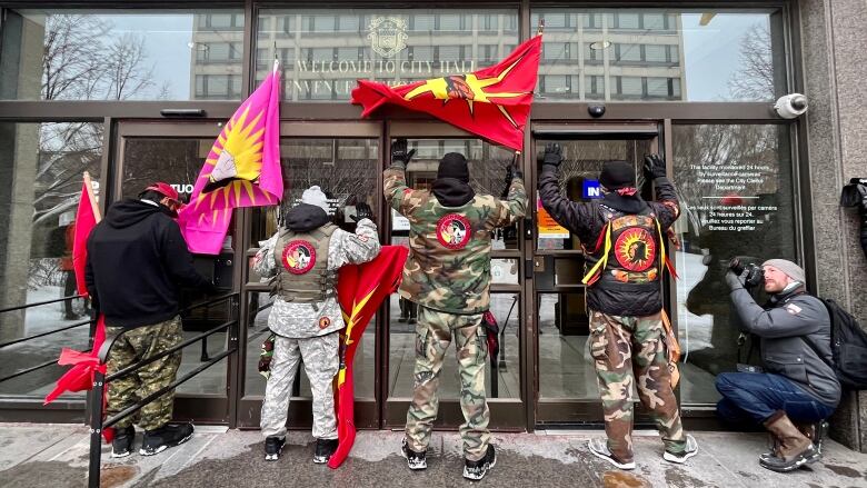 The backs of four people carrying protest flags and wearing camouflage are visible as they stand in front of the doors to the council building