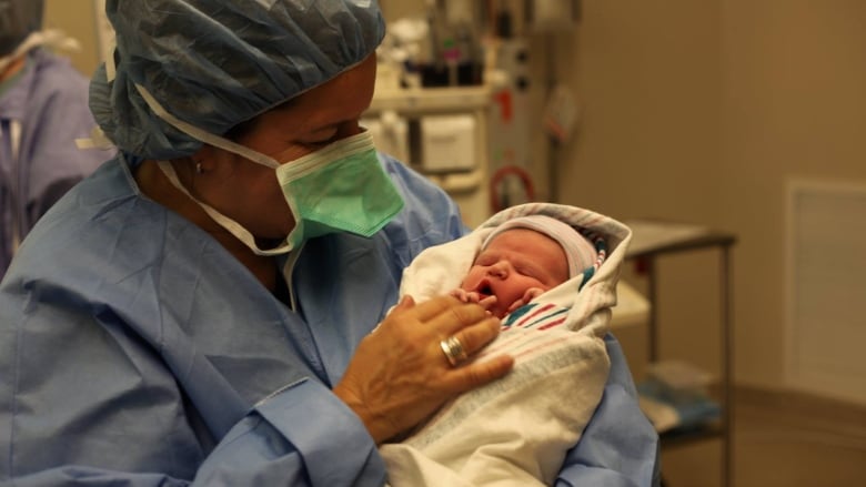 Woman in blue medical scrubs and medical mask holds a newborn baby. 