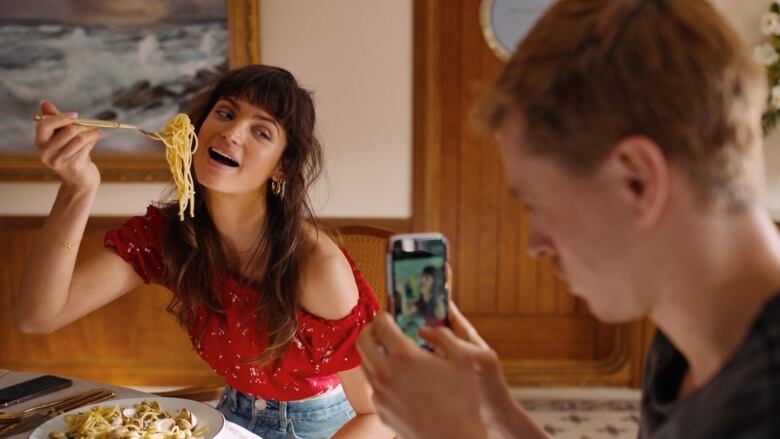 A woman with long dark hair holds up a forkful of spaghetti as she smiles for a photo being taken by a man with a smartphone.