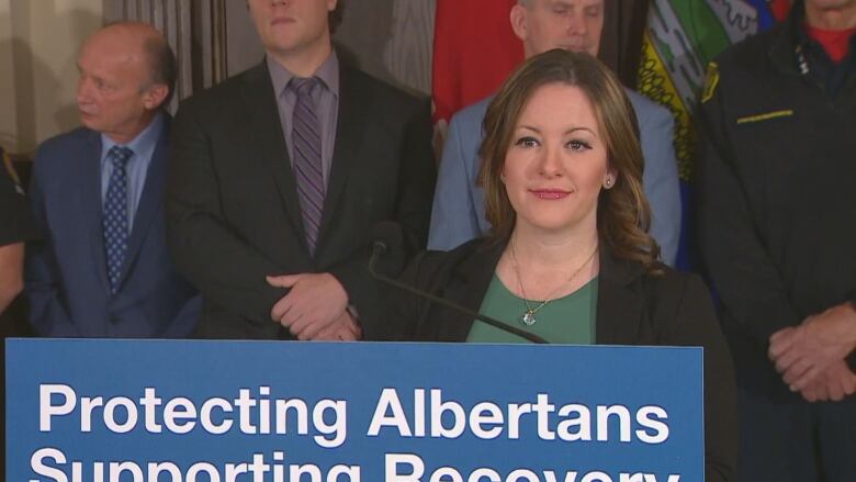 A woman wearing a blazer stands at a podium while three men in suits stand behind her.