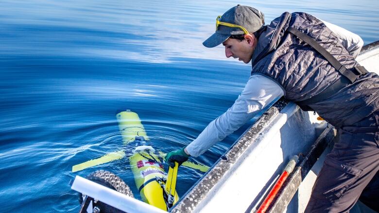 Man leans over the side of a small speedboat to touch the fin of a yellow underwater drone sticking out of a calm ocean surface.