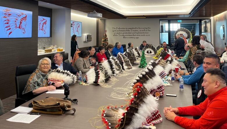 Several people sit at a long table in a conference room. Feathered headdresses sit on the table in front of many of the people.