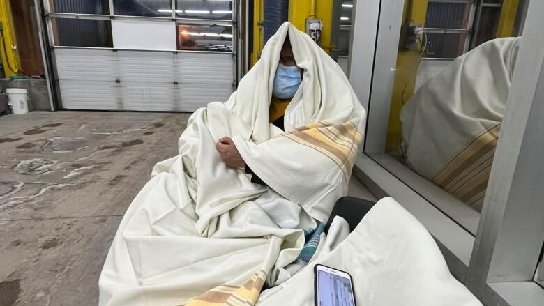 A woman sits wrapped in hospital blankets. She is wearing a blue medical face mask.