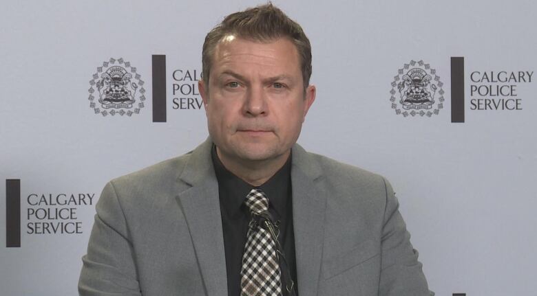 A middle-aged man with short brown hair and a clean-shaven face stands before a background that reads: 