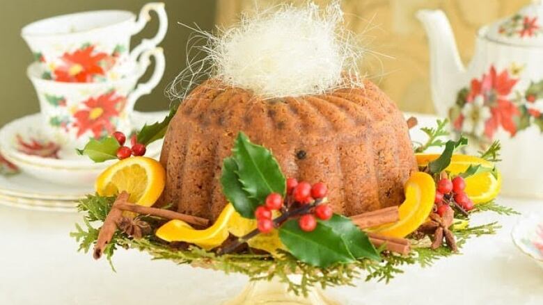 A cake in a bundt shape sits on a glass stand surrounded by lemons and decorative leaves.