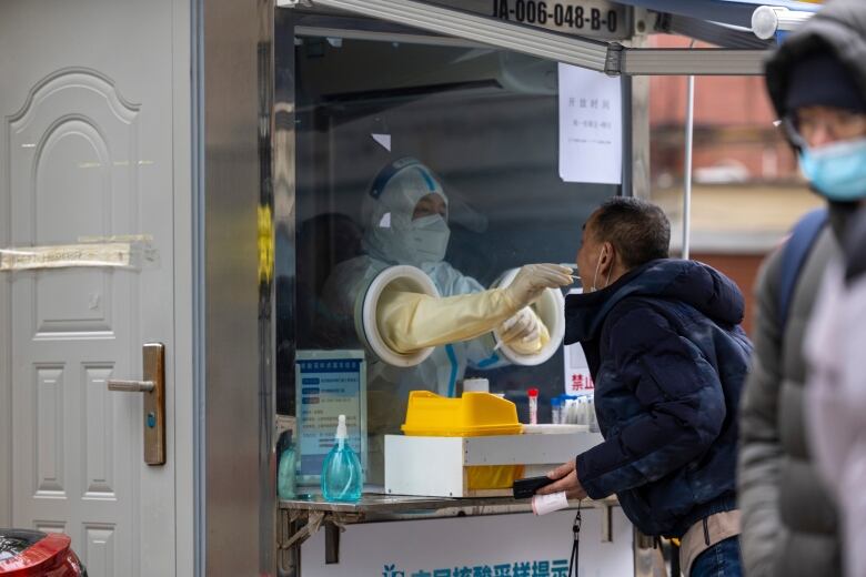 A healthcare worker in full personal protective equipment stands behind a glass barrier with two circular openings, allowing them to swab a man's mouth.