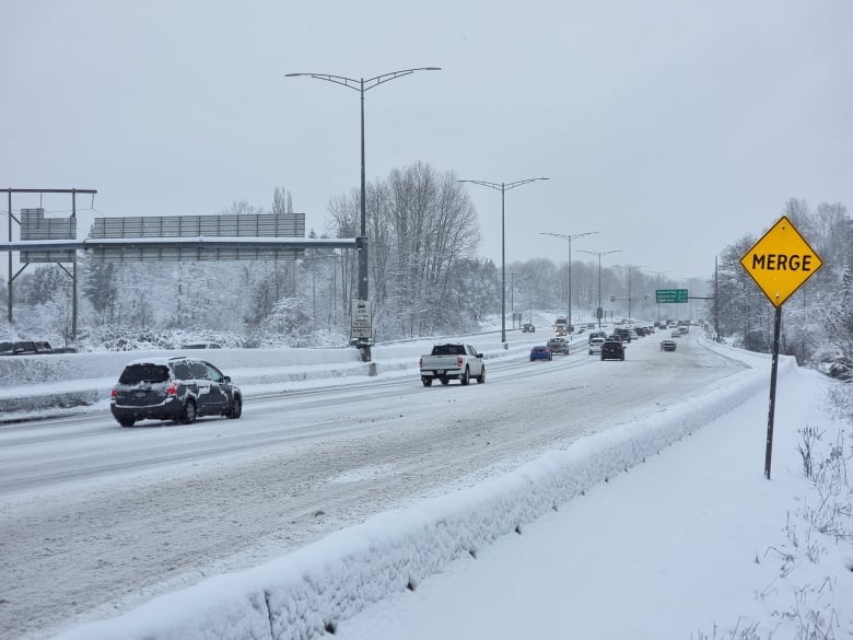 A row of vehicles on a snowy highway, with the guard rails covered by snow.