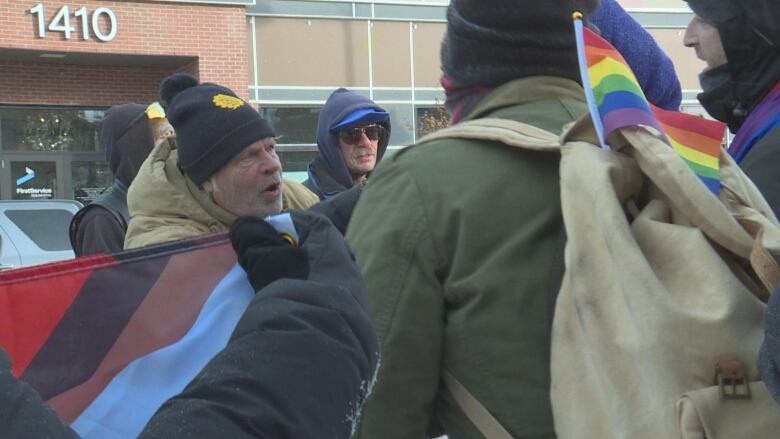 A man in a blue winter hat and a beige jacket yells at three people carrying LGBTQ flags, one of which yells back. 