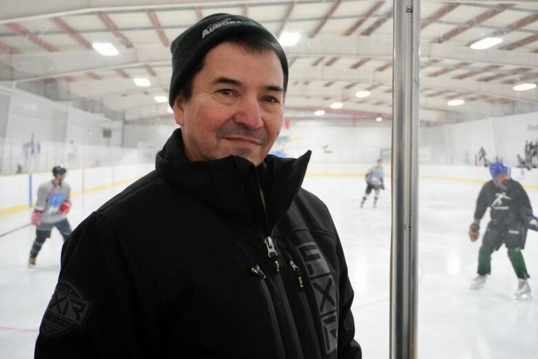 Man in black jacket stands in front of hockey rink, looking at the camera.